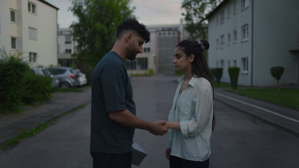 Still aus dem Film ELAHA. Ein Mann und eine Frau stehen auf einer Straße. Sie stehen sich gegenüber und er hält ihre Hand.