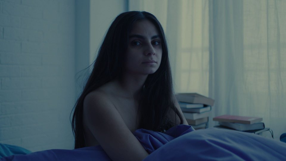 Stil aus dem Film ELAHA. Eine weiblich gelesene, weiße Person mit schwarzen, langen Haaren sitzt im Bett. Ihr nackter Oberkörper wird von einer dunkelblauen Bettdecke bedeckt.
