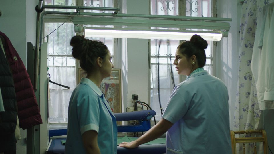Still aus dem Film ELAHA. Zwei Frauen stehen vor einem industriellen Bügelbrett, beide haben einen Kittel an. Sie scheinen gemeinsam zu diskutieren.
