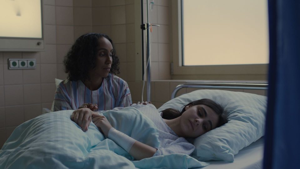 Still aus dem Film ELAHA. Eine junge Frau liegt schlafend in einem Krankenhausbett. Eine andere Frau sitzt neben dem Bett und hält die Hand der liegenden Frau.