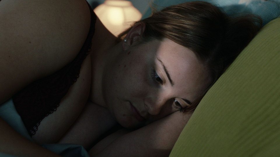Stil aus dem Film WAS WIR WOLLEN. Eine weiblich gelesene, weiße Person mit rot-braunem Haar und Sommersprossen liegt im schwarzen Spitzen-BH auf einem grünen Kissen.