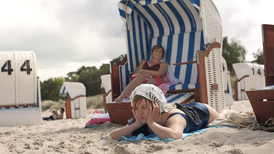 Still aus dem Film SPRICH MIT MIR. Eine junge Frau liegt auf einem blauen Handtuch im Badeanzug im Sand. Ihre Hände stützend ihren Kopf, sie scheint genervt zu sein. Im Hintergrund befinden sich mehrere Strandkörbe, in einem sitzt eine ältere Frau ebenfalls in Badekleidung.