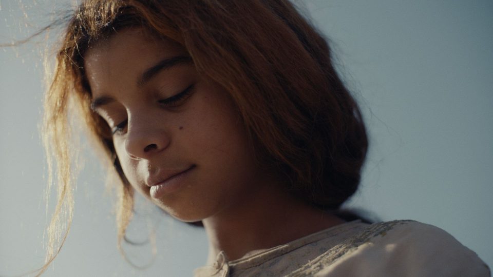 Still aus dem Film A CALLING . FROM THE DESERT . TO THE SEA. Portrait eines jungen Mädchens, das nach unten sieht. Ihr Haar weht im Wind und wird von der Sonne angeleuchtet.