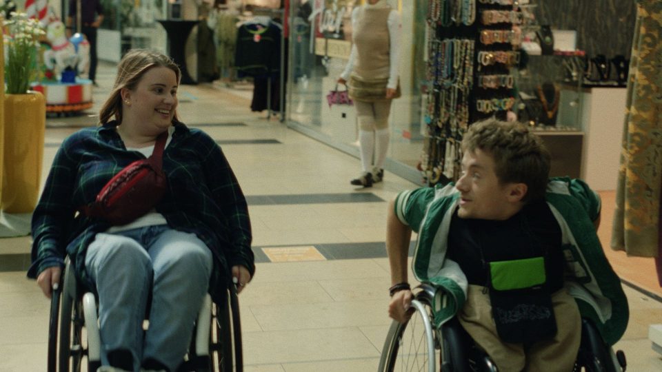 Still aus dem Film WAS WIR WOLLEN. Eine Frau und ein Mann fahren sitzend im Rollstuhl durch ein Kaufhaus. Beide schauen sich lächelnd an.