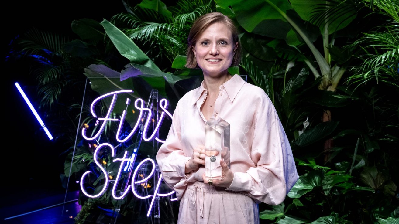 Die Preisträgerin Clara Gerst hält den First Steps Award für die Kategorie "No Fear Award" in ihren Händen und steht vor großen Pflanzen, sowie einer lila Leuchtschrift mit der Aufschrift "First Steps". Clara Gerst hat kurze, glatte, hellbraune Haare und trägt einen rosa Hosenanzug.