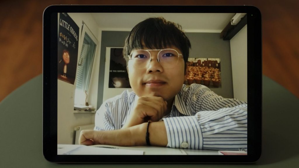 Nominierter Hao Yu. Junge, asiatisch und männlich gelesene Person mit schwarzen Haaren sitzt am Schreibtisch und stützt sein Kinn auf seine rechte Hand. Er trägt eine silberne Brille und ein blau-weiß gestreiftes Hemd.